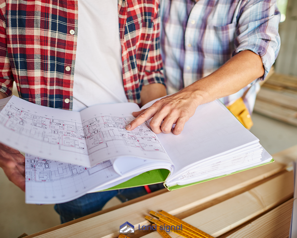 Two builders reviewing detailed architectural floor plans on a construction site, with one pointing to specific measurements on the blueprint.